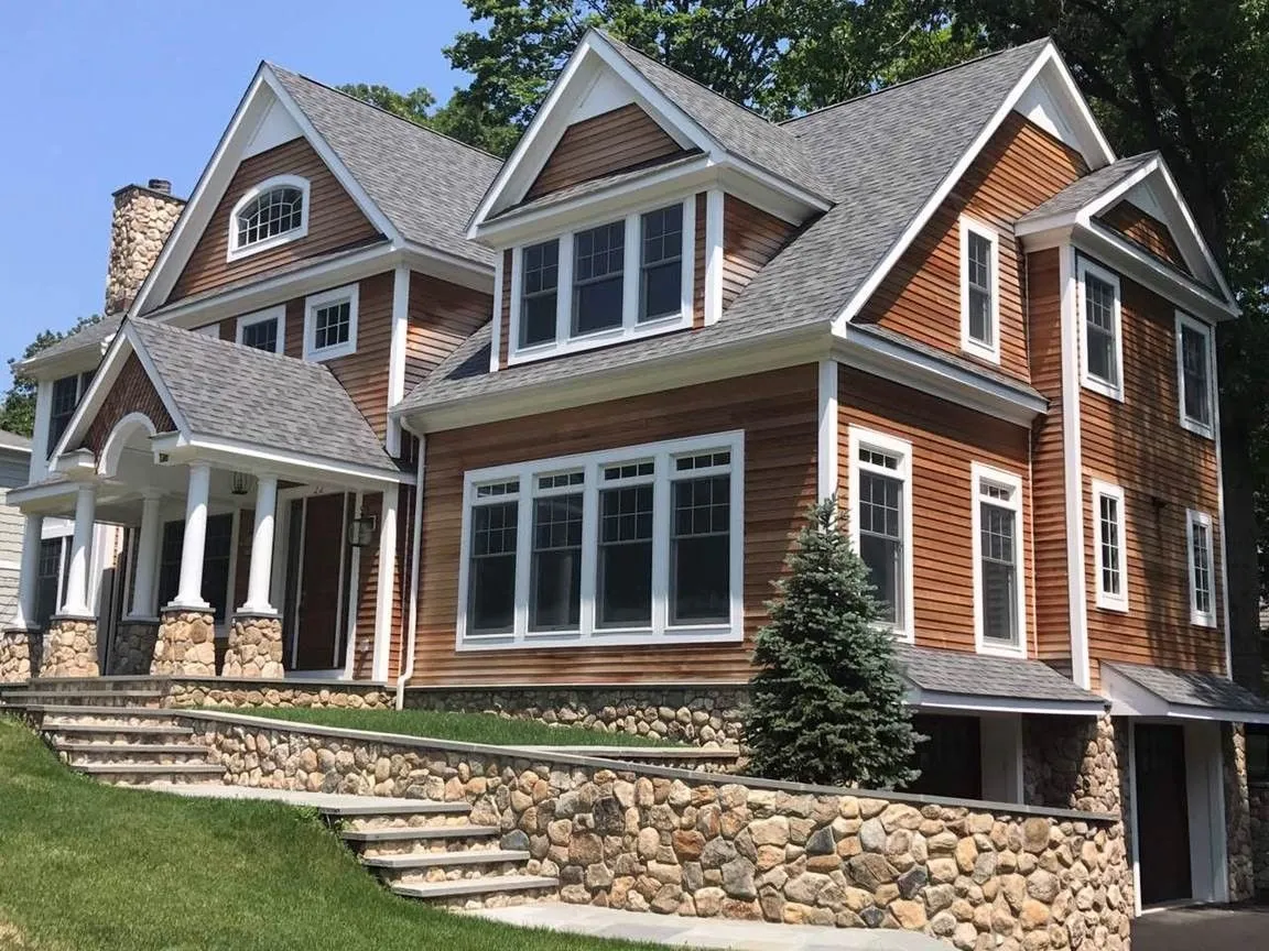 Large brick and wood house with stone steps, wraparound porch, and gabled roof on a grassy slope