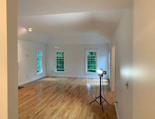 Empty room with hardwood floors, white walls, three windows, and a light on a tripod stand.