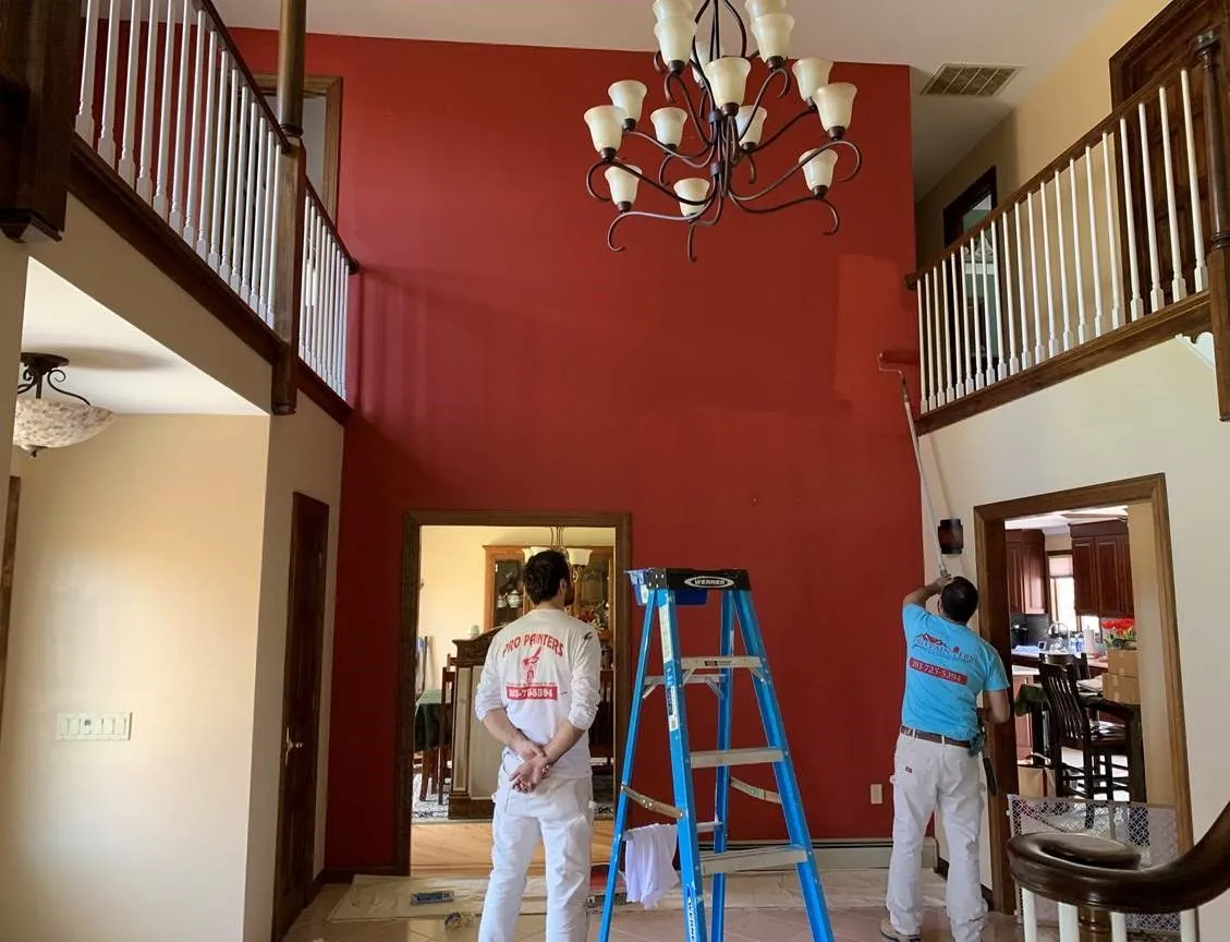 Two workers painting a red-walled foyer, with a blue ladder and chandelier overhead.